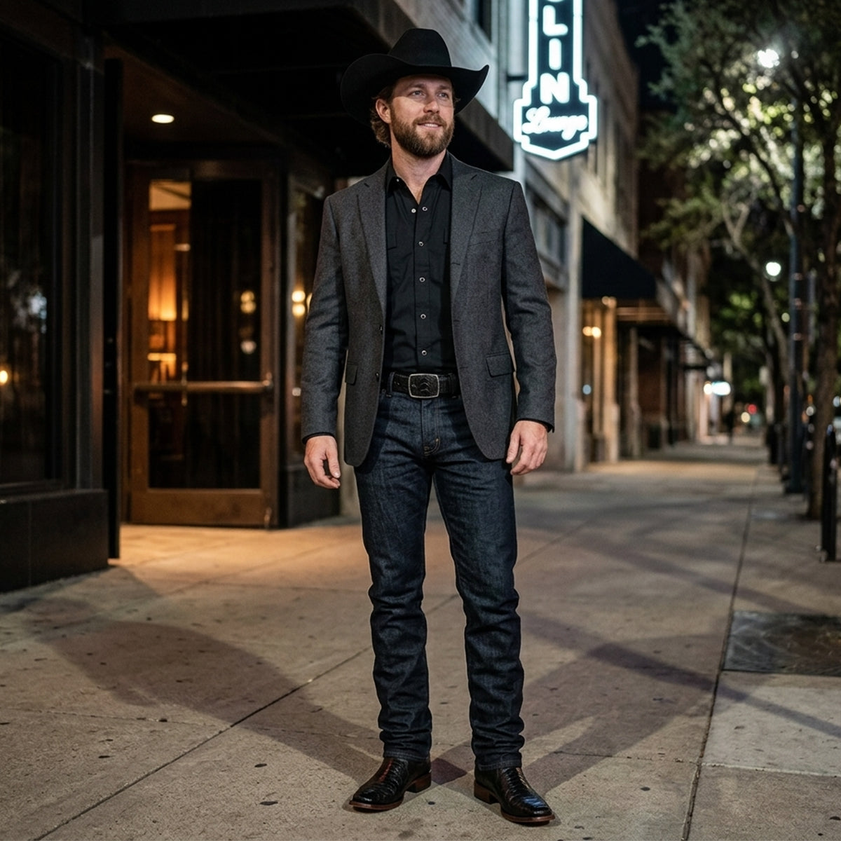 A man wearing a dark grey blazer jacket over a black western snap shirt, dark wash blue jeans, a black leather belt, black ostrich leg cowboy boots, and a black cowboy hat standing on a city street at night.