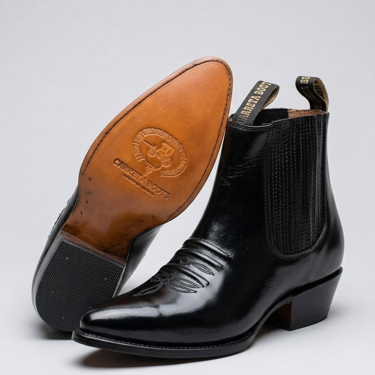 A pair of black leather ankle boots shown on a light gray studio background, with one boot standing upright and the other tilted to display the tan outsole. The outsole is embossed with the brand name 'CARRETA BOOTS'.