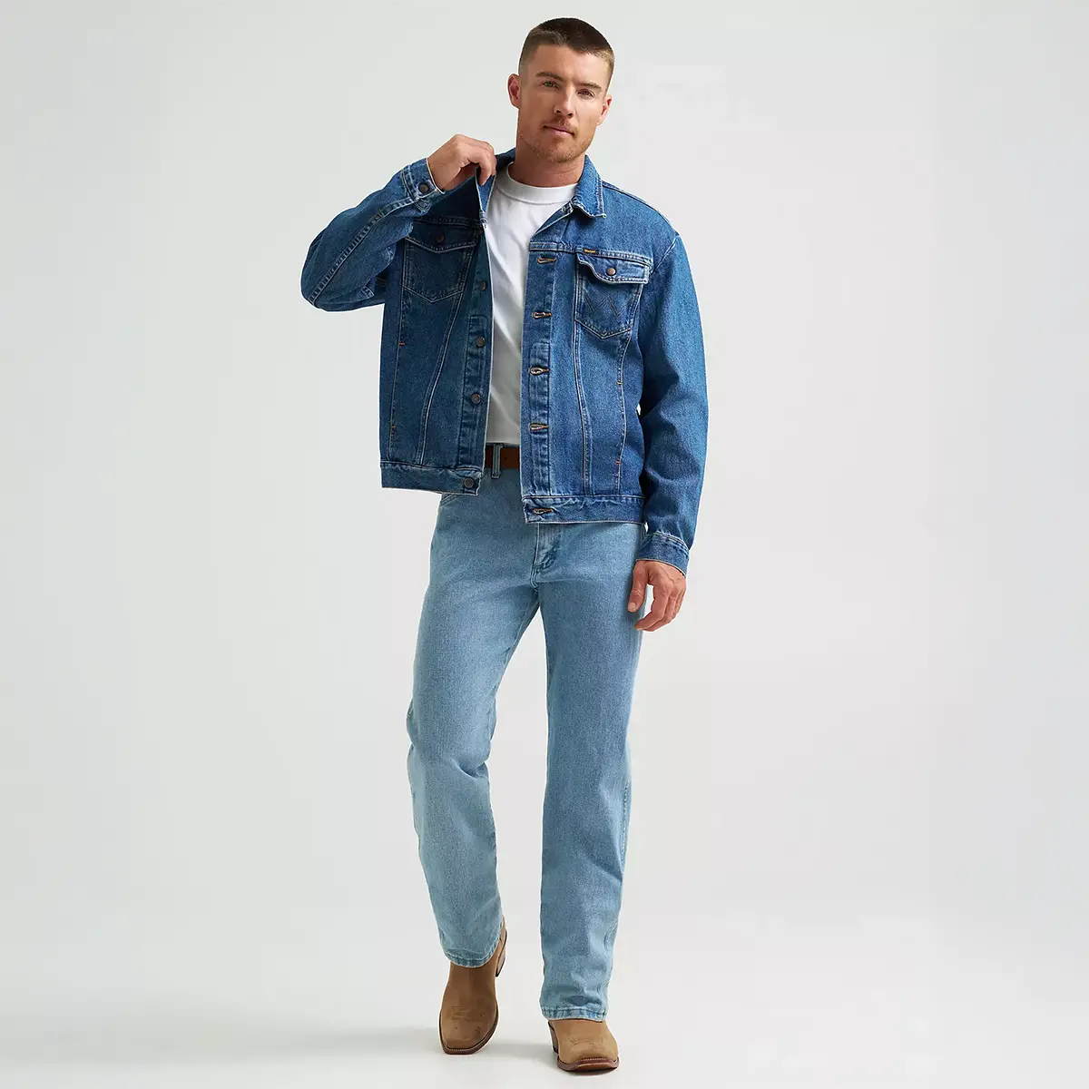 Full-length lifestyle shot of the model wearing the Wrangler unlined stonewash denim jacket with light wash jeans and brown leather Western boots.