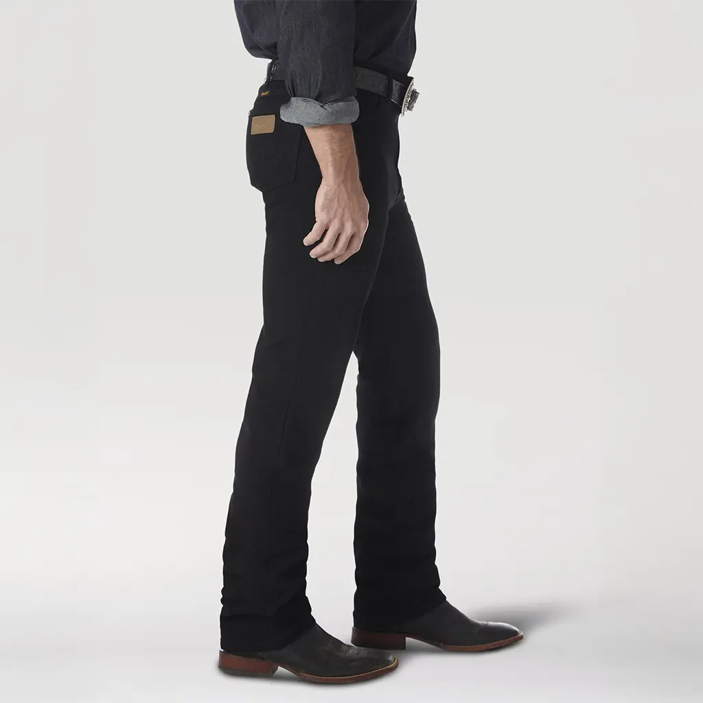Side profile view of a model wearing the Wrangler 0936WBK Shadow Black jeans showcasing the slim leg fit over leather boots.