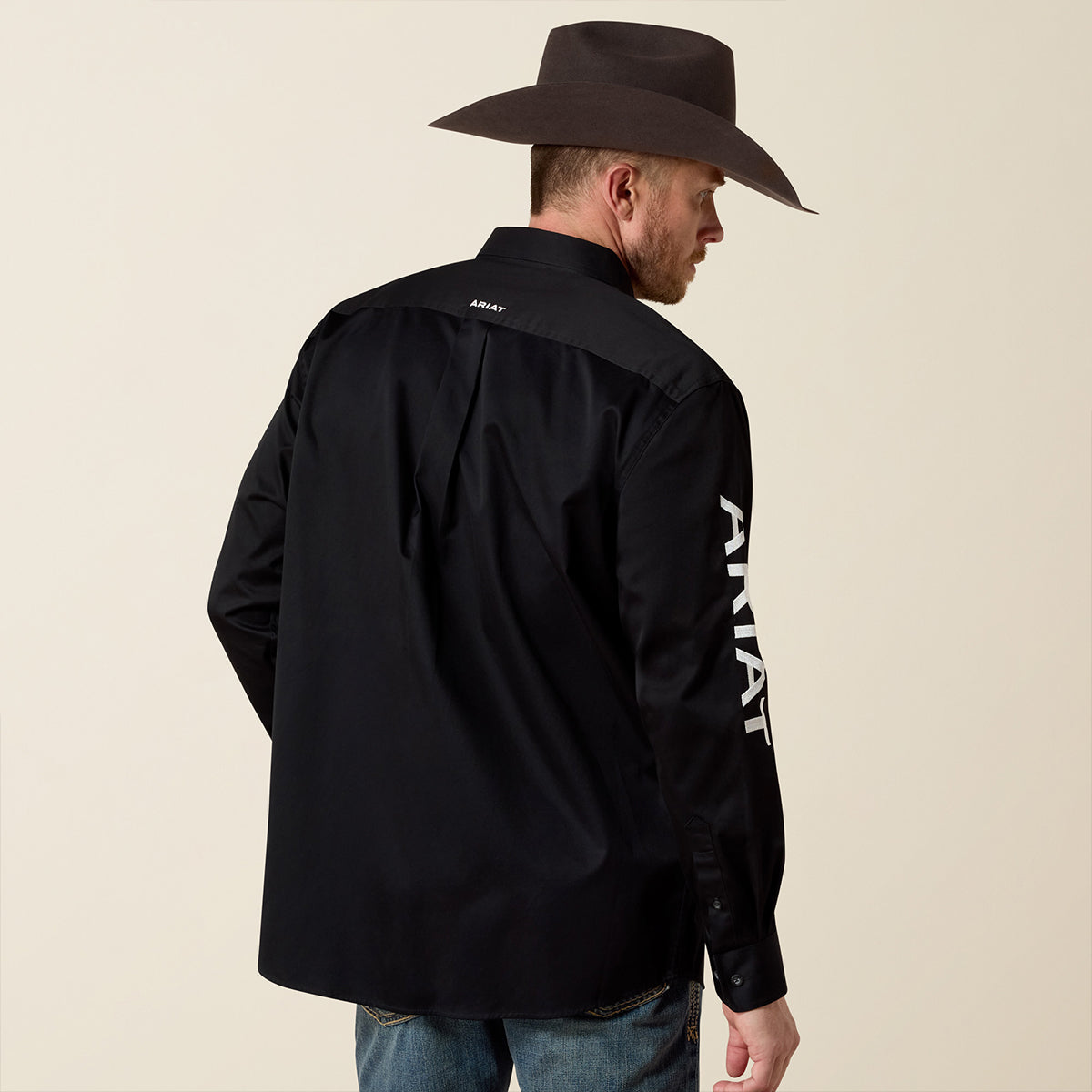 Man wearing a black shirt with Ariat branding on the back and side, standing against a beige background