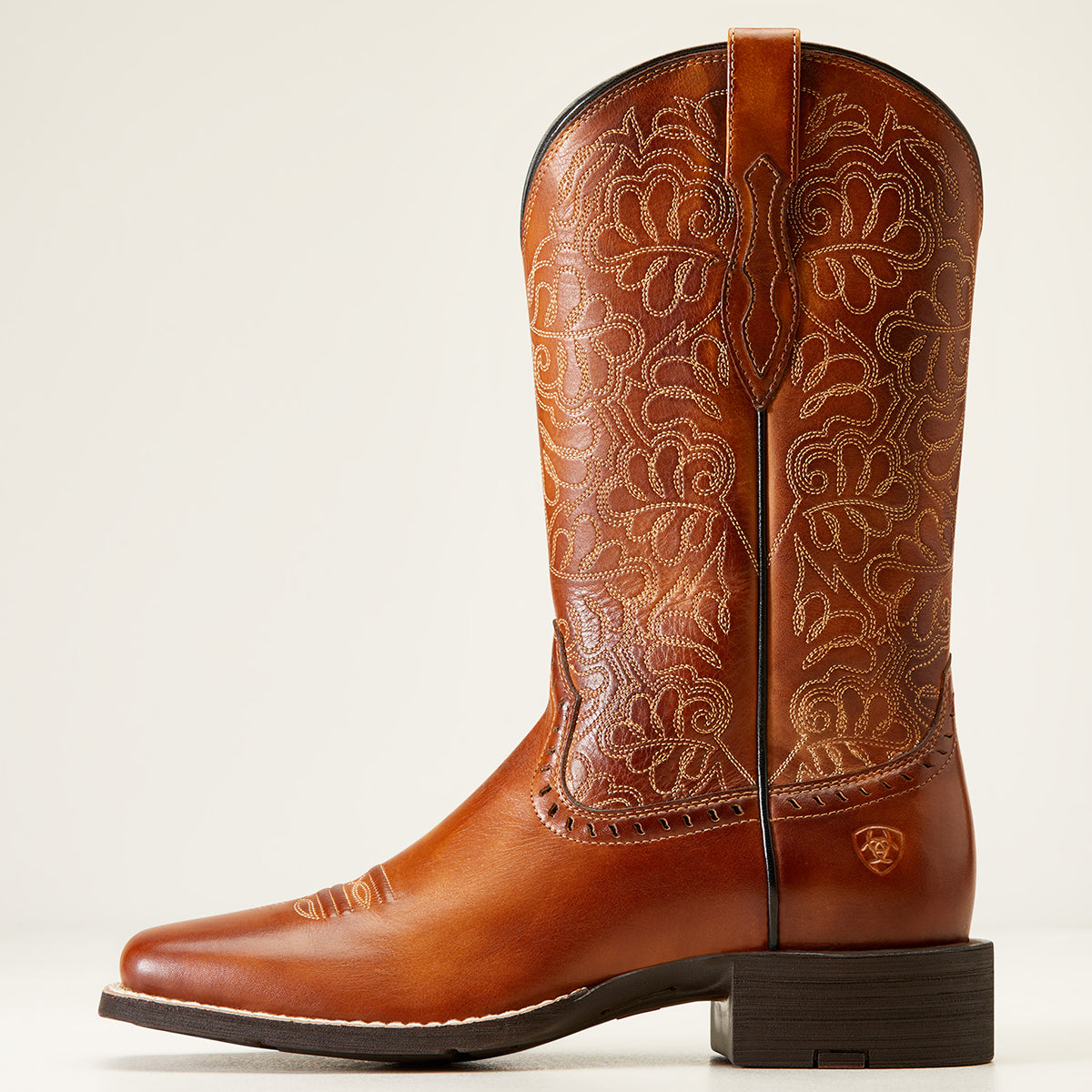 A single brown leather Ariat cowgirl boot with intricate floral tooled embroidery on the shaft, a square toe, and a dark stacked heel, shown against a plain light background.