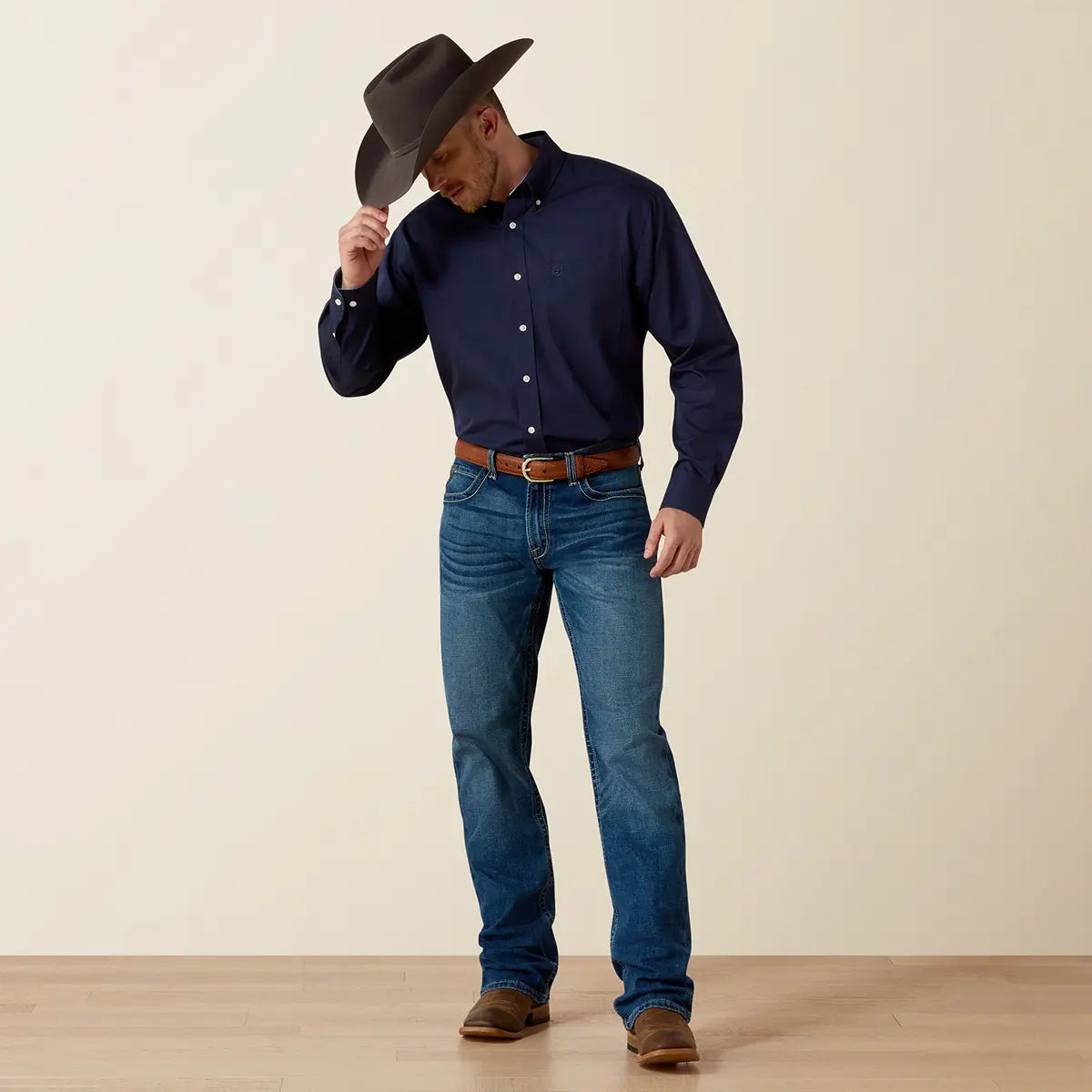 Full-length lifestyle view of the model adjusting his dark cowboy hat while wearing the Ariat navy blue wrinkle-free shirt with denim jeans and leather boots.