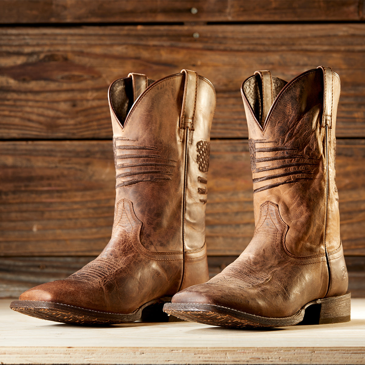 Ariat men's brown leather cowboy boots with western stitching and an American flag motif on the shaft, displayed on a light wooden surface against a rustic dark wood plank background.