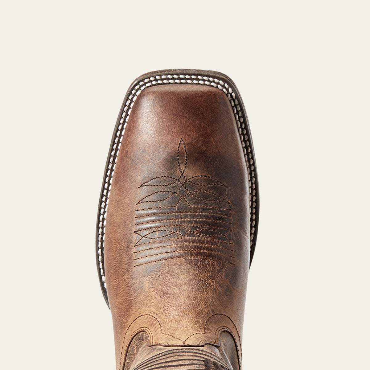 A close-up top-down view of a brown leather cowboy boot square toe, featuring decorative stitching and a square vamp on a light beige background.
