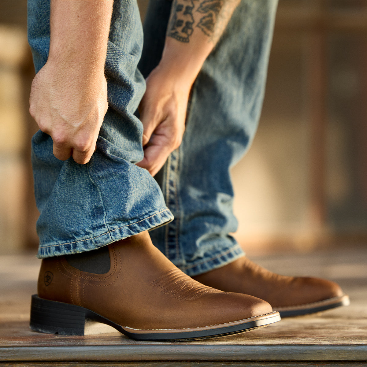 A close-up of a person wearing brown leather Ariat cowboy boots with blue jeans, adjusting the hem of the jeans. The boots feature a low block heel, a side elastic gusset, and a stitched toe.