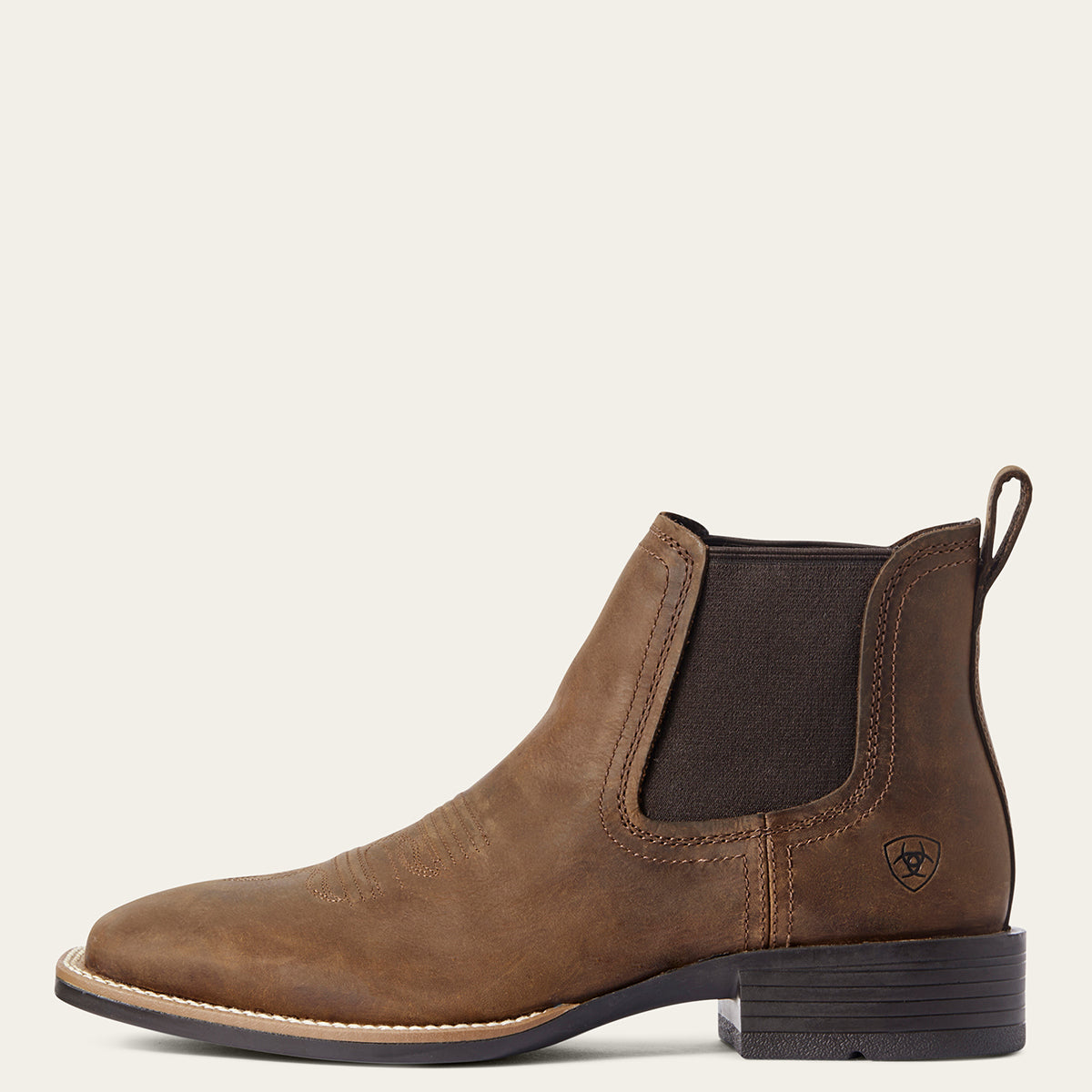 Ariat men's brown leather Chelsea-style ankle boot with a dark brown elastic side panel and a low black heel, shown in profile against a light cream background.