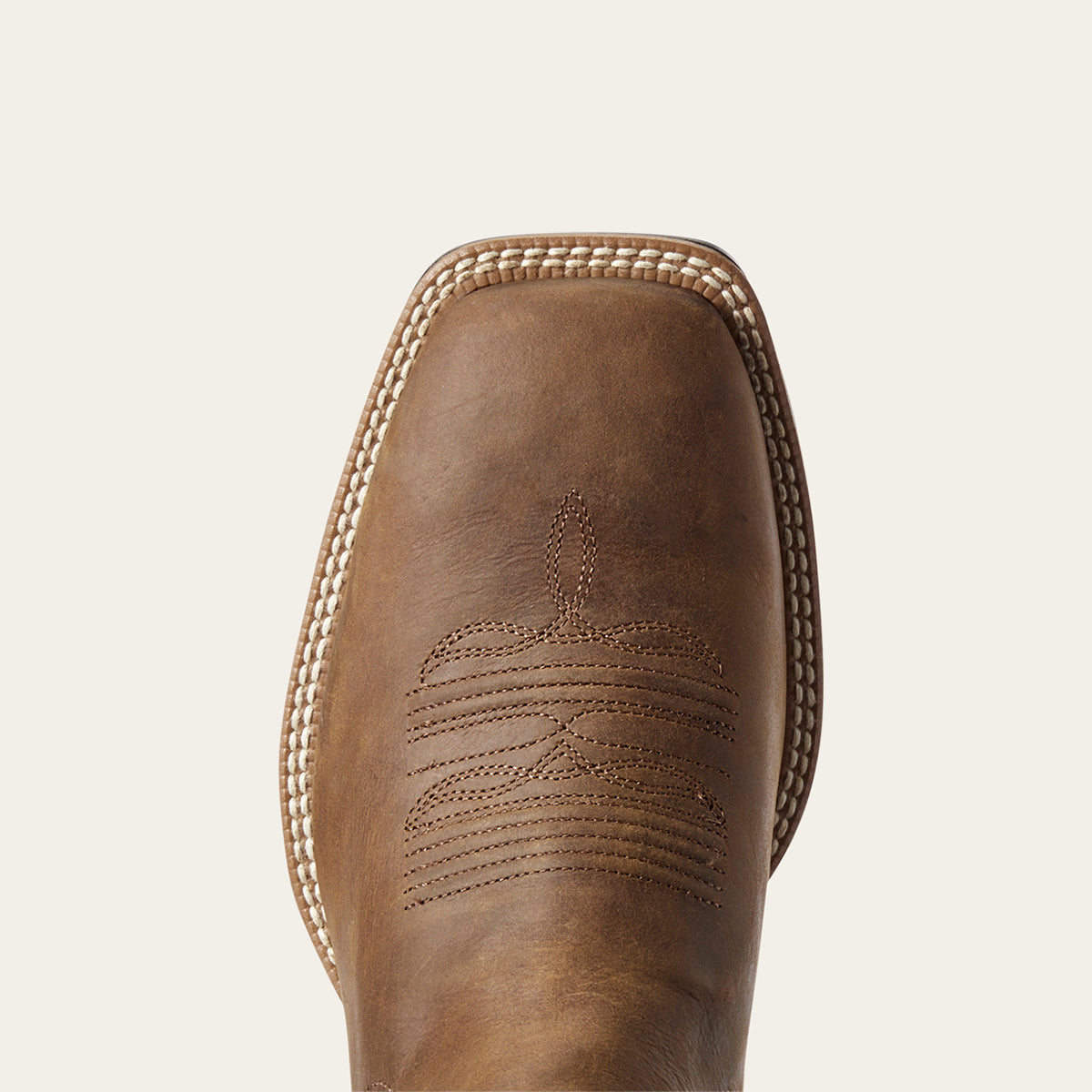 Close-up of the Ariat toe of a brown leather cowboy boot with decorative stitching and contrast welt stitching, shown against a light beige background.