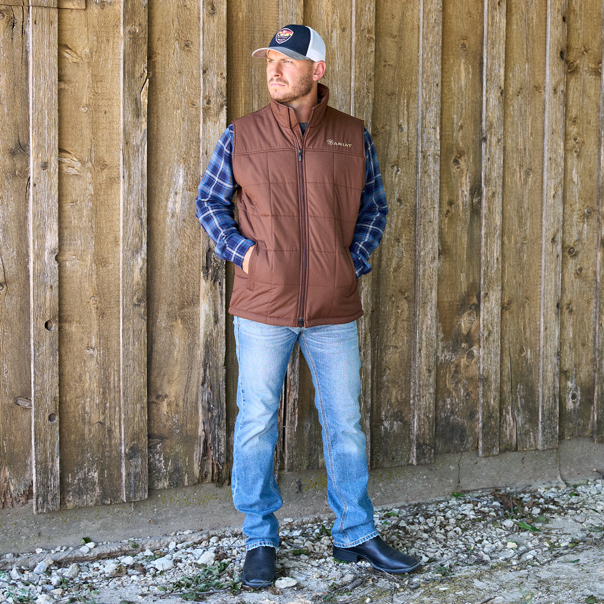 A man wearing a brown quilted zip-front vest over a blue plaid shirt, blue jeans, and black Ariat boots stands in front of a rustic wooden wall.