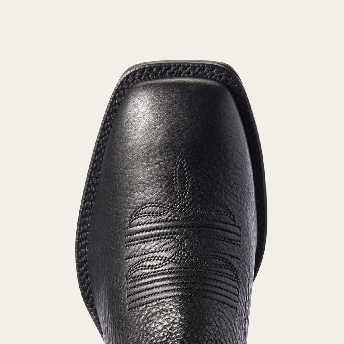 Close-up of Ariat black leather cowboy boot square toe showing the textured grain, stitched edge, and western-style embossed stitching on a light off-white background.