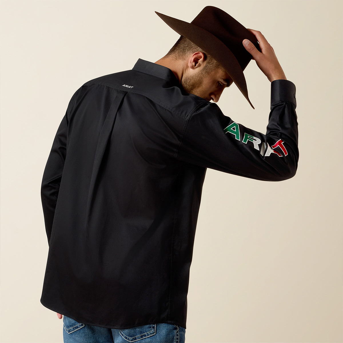 Man wearing a black Ariat shirt with colorful text on sleeves and a brown hat, against a beige background