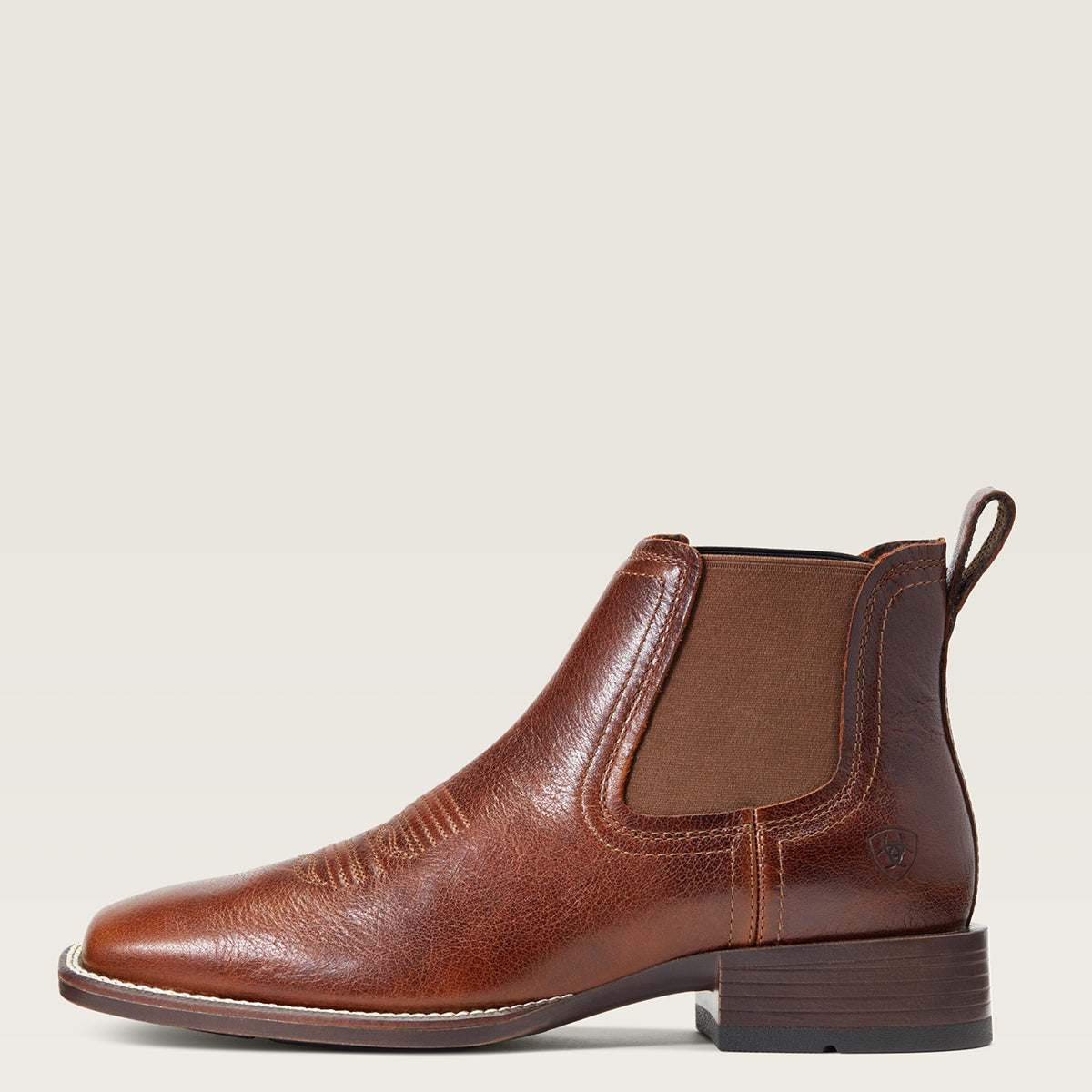 Ariat men's brown leather Chelsea boot with a low block heel, featuring contrast stitching, elastic side panels, and a pull tab, shown in profile against a light beige background.