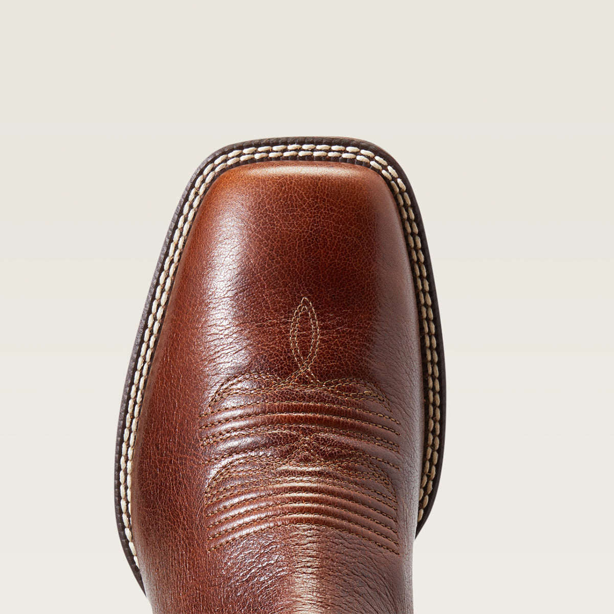 Close-up of the Ariat square toe of a brown leather cowboy boot with decorative stitching and a square toe, shown against a light beige background.