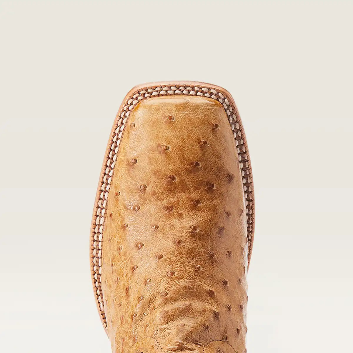 Close-up of Ariat tan ostrich leather boot corner with decorative whipstitching, set against a light beige background.