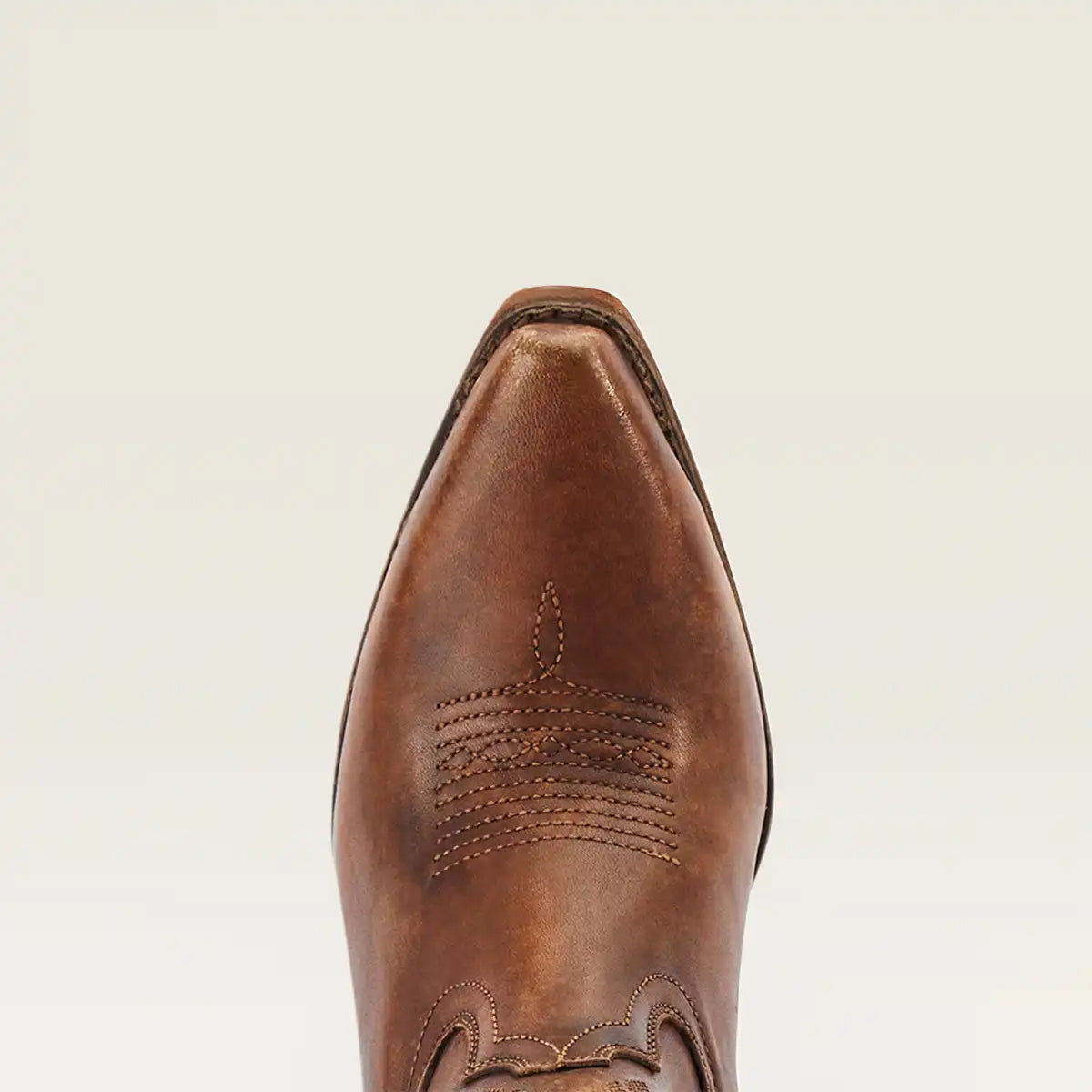 A close-up top-down view of a brown leather cowgirl boot toe, showcasing pointed styling, decorative stitching, and a distressed finish against a light off-white background.