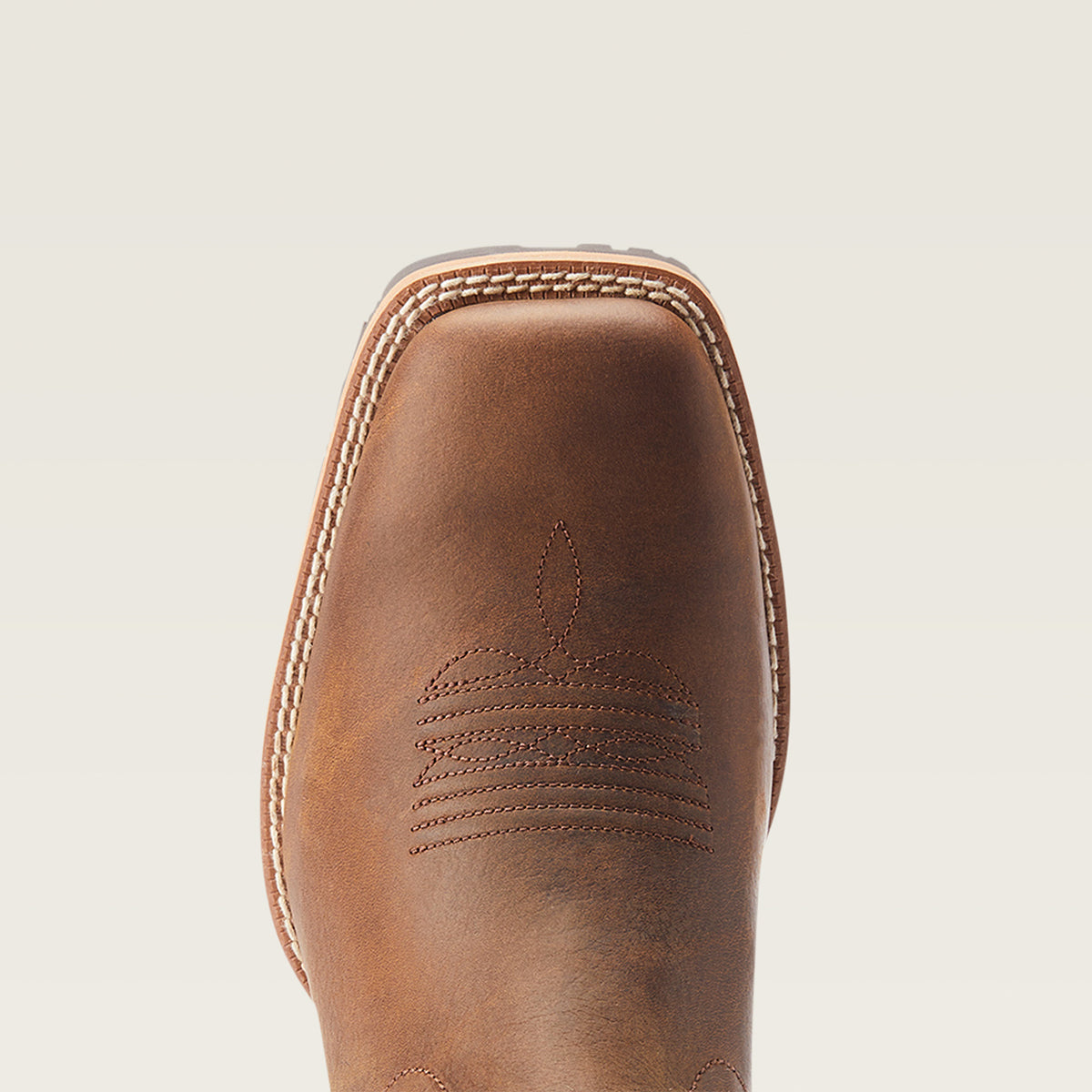 Close-up of the Ariat square toe of a brown leather cowboy boot, showing decorative stitching and contrast stitching along the edge, on a light beige background.