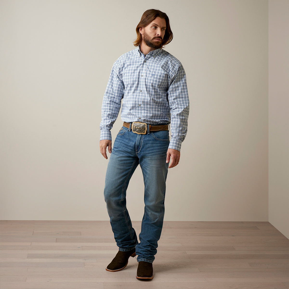 Full-body view of a man wearing the Ariat Men's Pro Series Othman blue and white plaid long sleeve button-down shirt paired with medium-wash jeans, brown cowboy boots, and a trophy belt buckle.