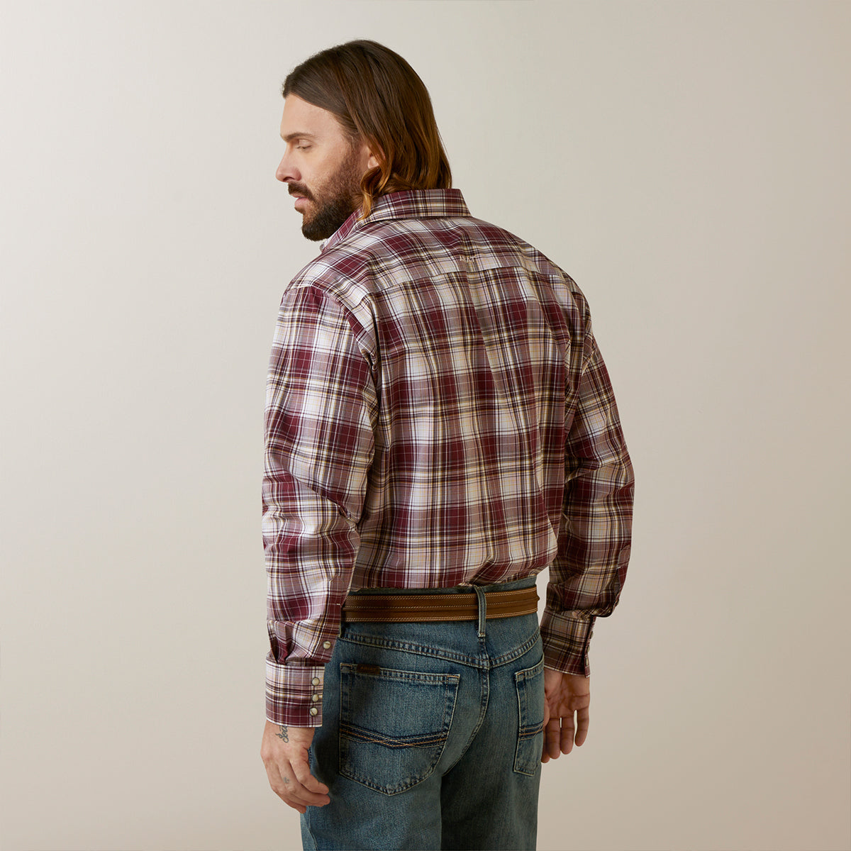 A man with long brown hair and a beard, shown from the back wearing a long-sleeve plaid button-down shirt in burgundy, beige, and white, paired with blue jeans and a brown belt, against a plain light beige background.
