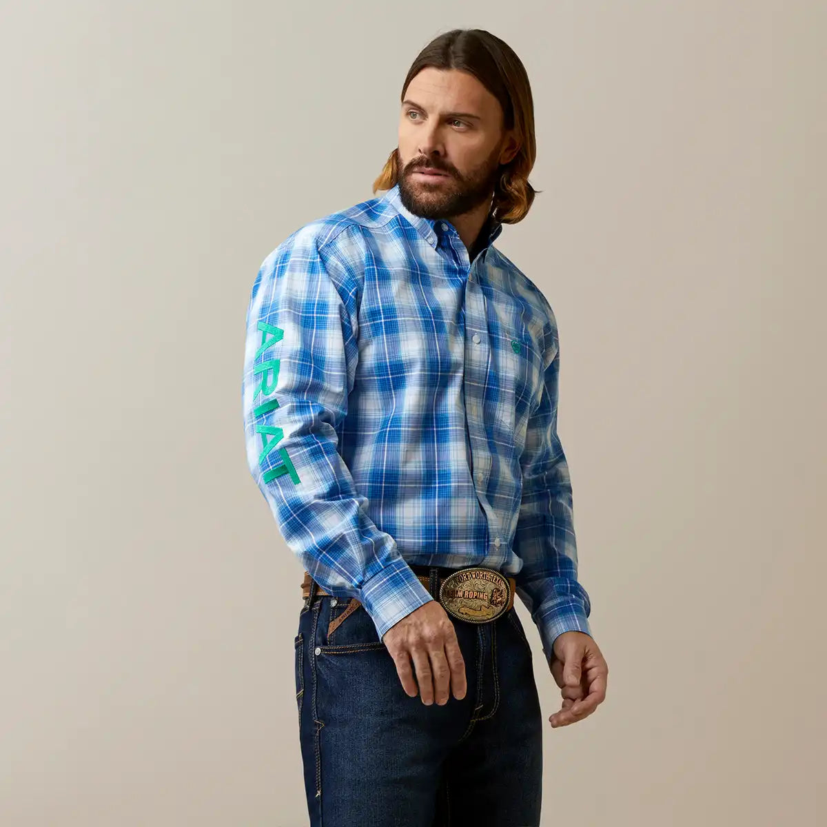A man wearing a blue and white plaid western long-sleeve button-down shirt with green 'ARIAT' branding printed vertically on the right sleeve, paired with dark blue jeans and a large western belt buckle, set against a plain beige studio background.