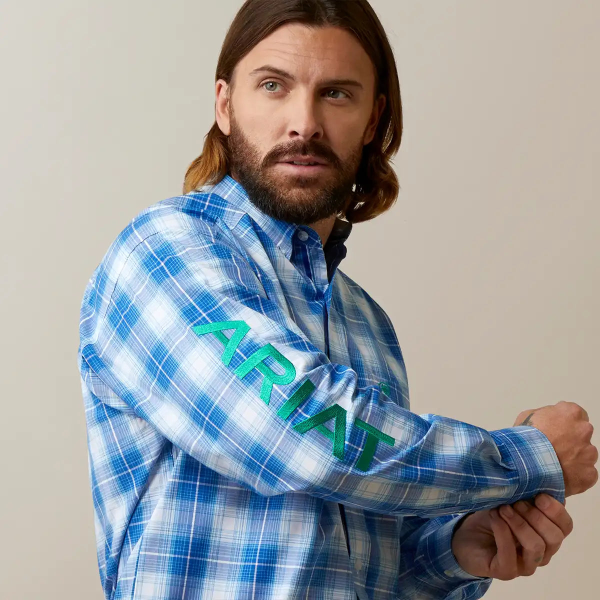 A man wearing a blue and white plaid long-sleeve button-up shirt with 'ARIAT' printed in green across the sleeve, posed against a plain beige studio background.