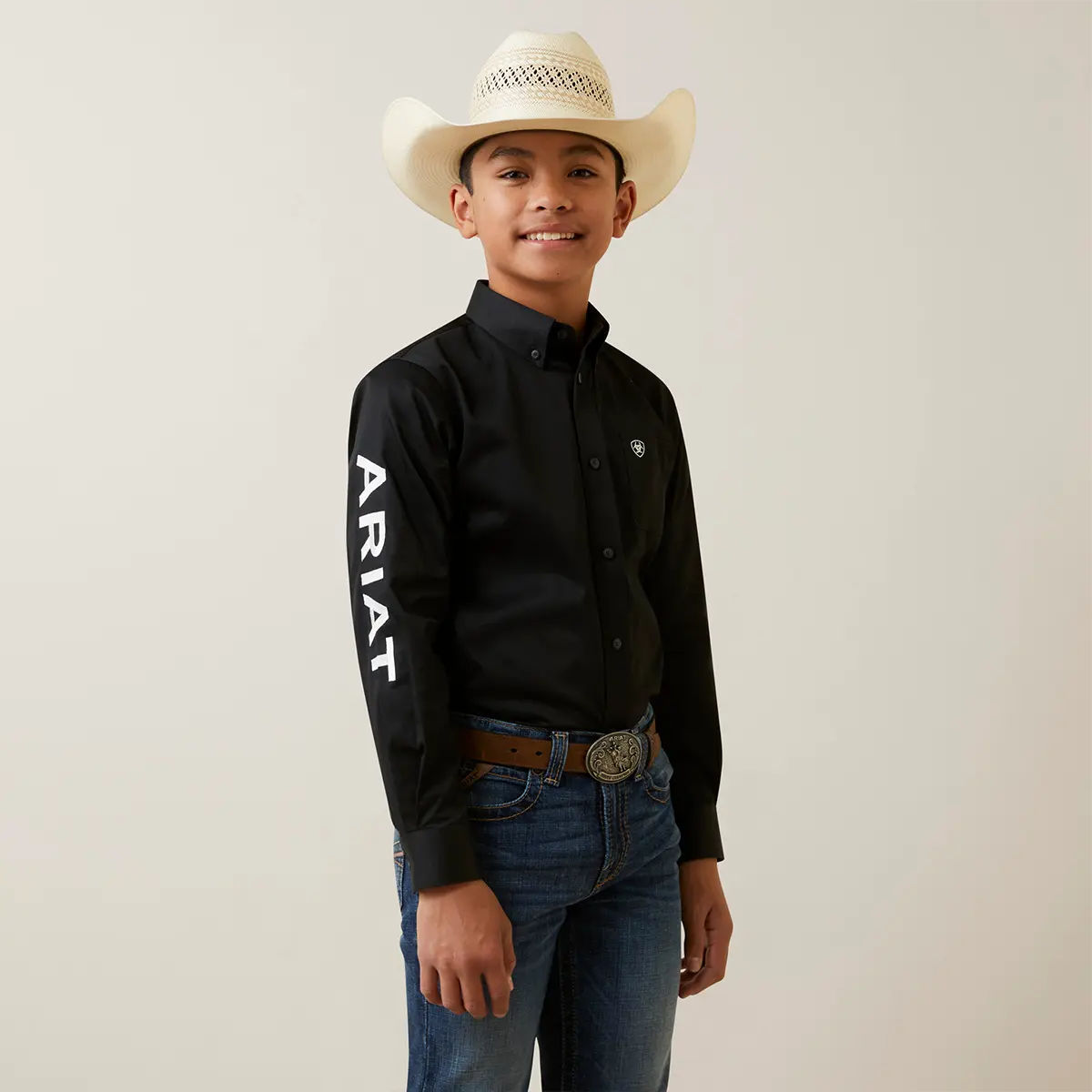 Front studio view of a young boy wearing the Ariat Boys' black solid twill team logo shirt with a straw cowboy hat and jeans.
