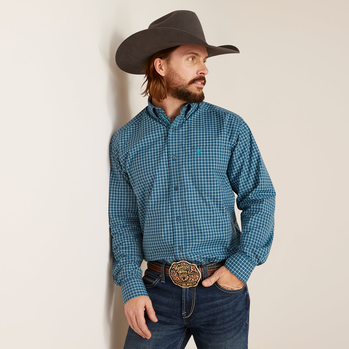 A man wearing a blue and teal checkered western long-sleeve button-down Ariat shirt, dark blue jeans, a brown cowboy hat, and a large gold buckle belt, standing against a plain off-white wall.