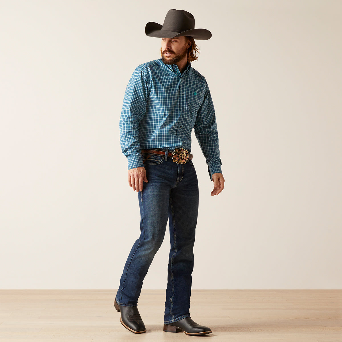 A man modeling a blue and black checkered long-sleeve western Ariat shirt, paired with dark blue jeans, a brown cowboy hat, and black boots against a neutral studio background.