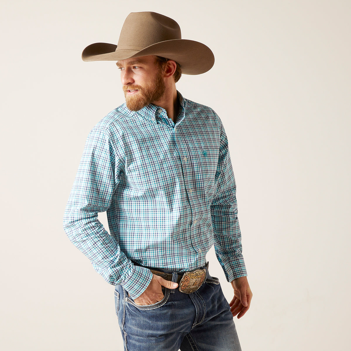 A man wearing a tan cowboy hat, a light blue and teal plaid western button-down Ariat shirt, and blue jeans, standing against a plain off-white studio background.