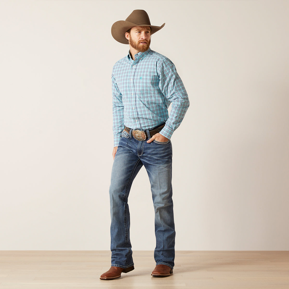 Man wearing a blue plaid Ariat shirt, jeans, and a cowboy hat on a plain background