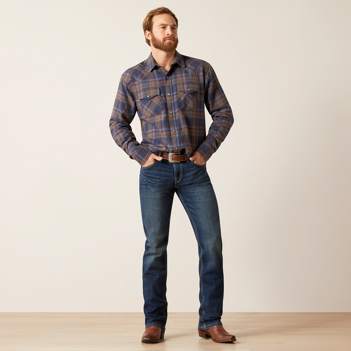 A man wearing a long-sleeve plaid western-style button-down Ariat shirt in blue, brown, and orange, paired with dark blue jeans and brown leather boots, standing against a plain beige studio background.