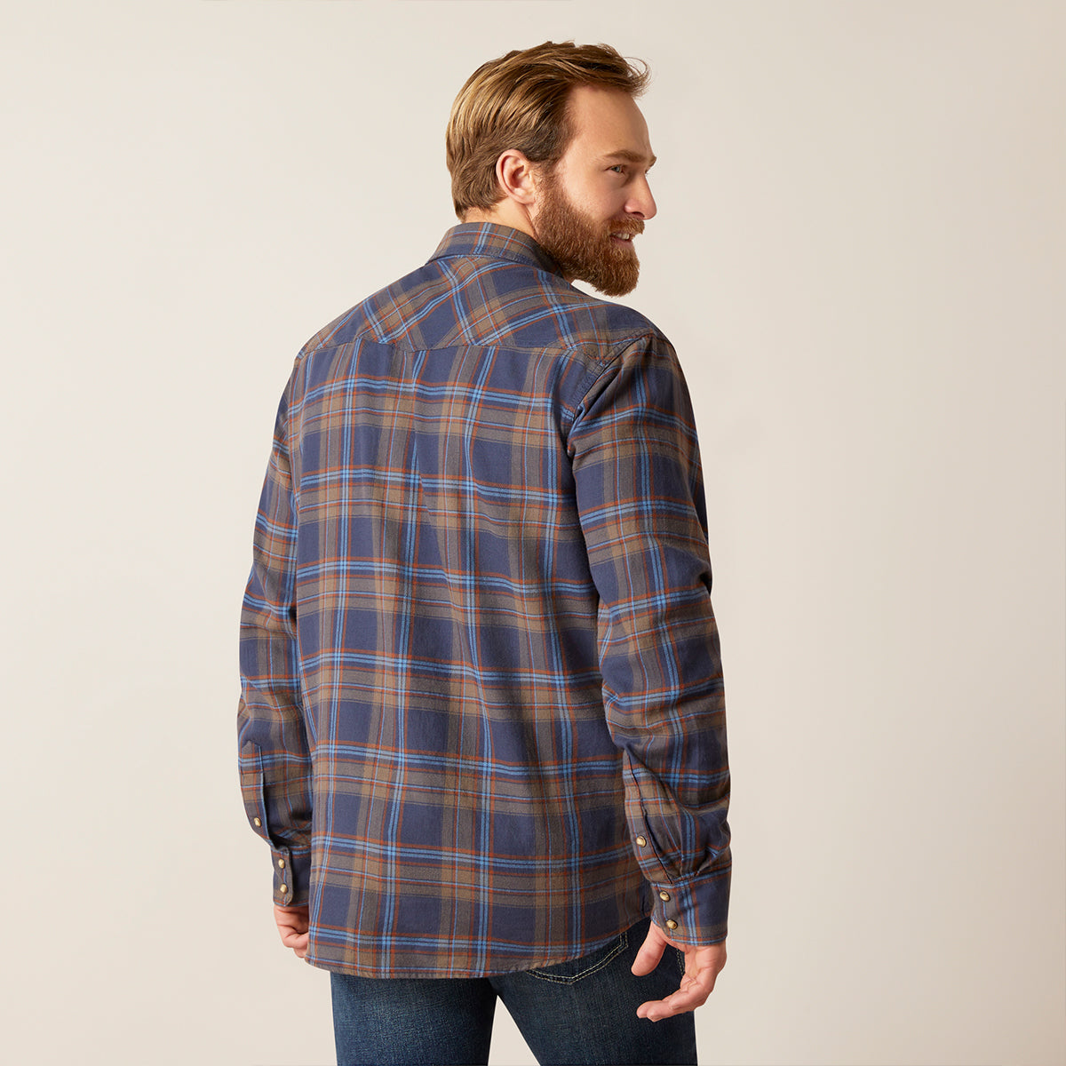 A man with a beard is shown from the back wearing a blue, brown, and tan plaid button-down Ariat shirt, paired with blue jeans, against a plain light beige background.