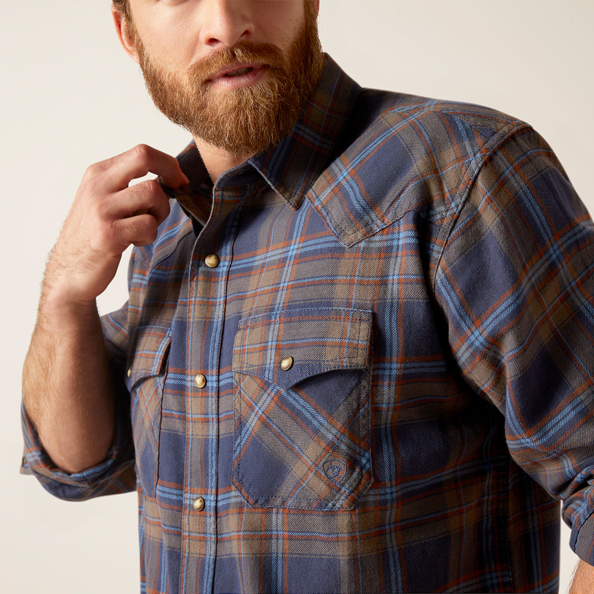 A close-up of a man wearing a blue, brown, and orange plaid button-down shirt, adjusting the collar with one hand.