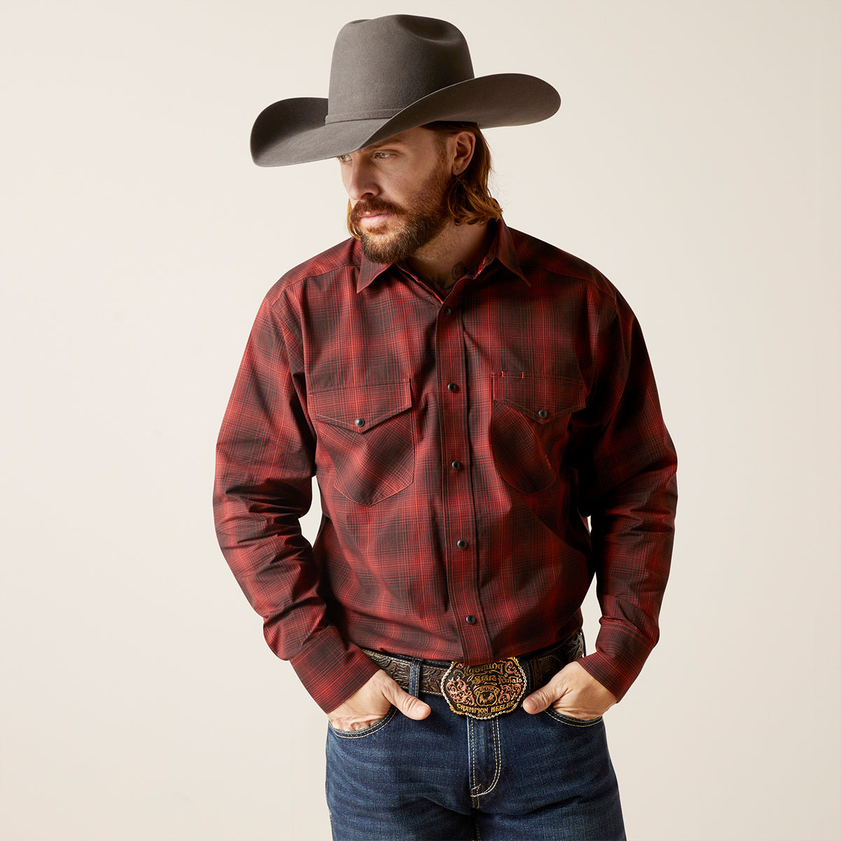 A man wearing a red and black plaid western long-sleeve button-up Ariat shirt, blue jeans, and a gray cowboy hat, standing against a light beige studio background.