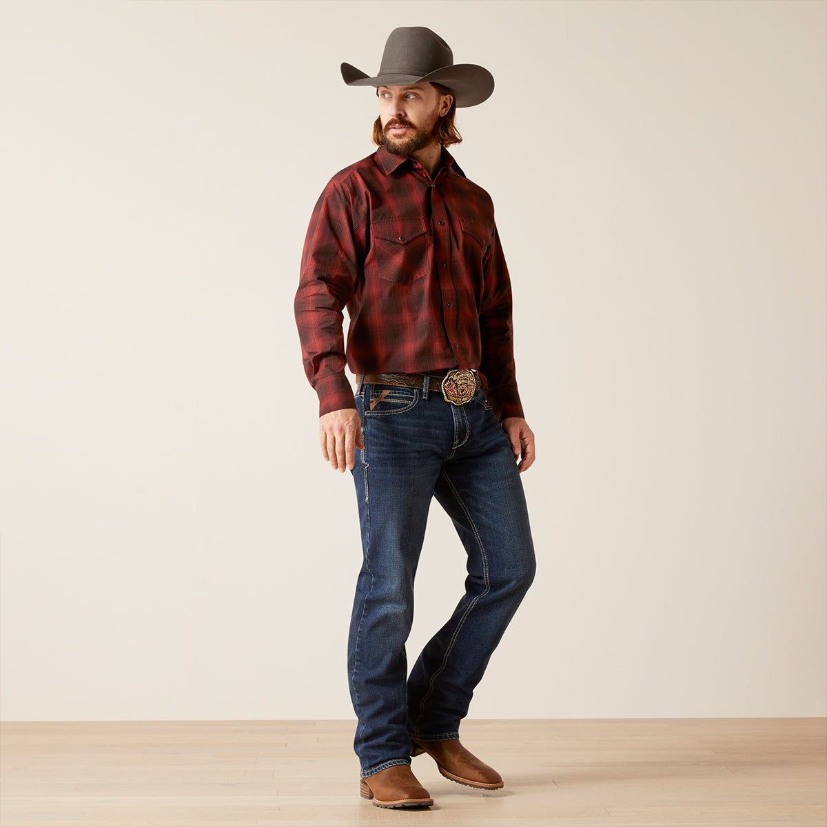 A man modeling a red and black buffalo plaid western long-sleeve Ariat shirt, paired with dark blue jeans, a brown cowboy hat, and brown leather boots against a neutral studio backdrop.