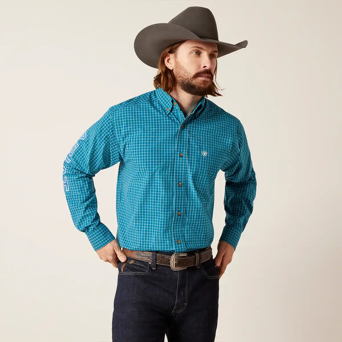 A man wearing a turquoise and blue checkered long-sleeve western Ariat shirt, dark jeans, a brown belt, and a dark brown cowboy hat, standing against a light beige background.