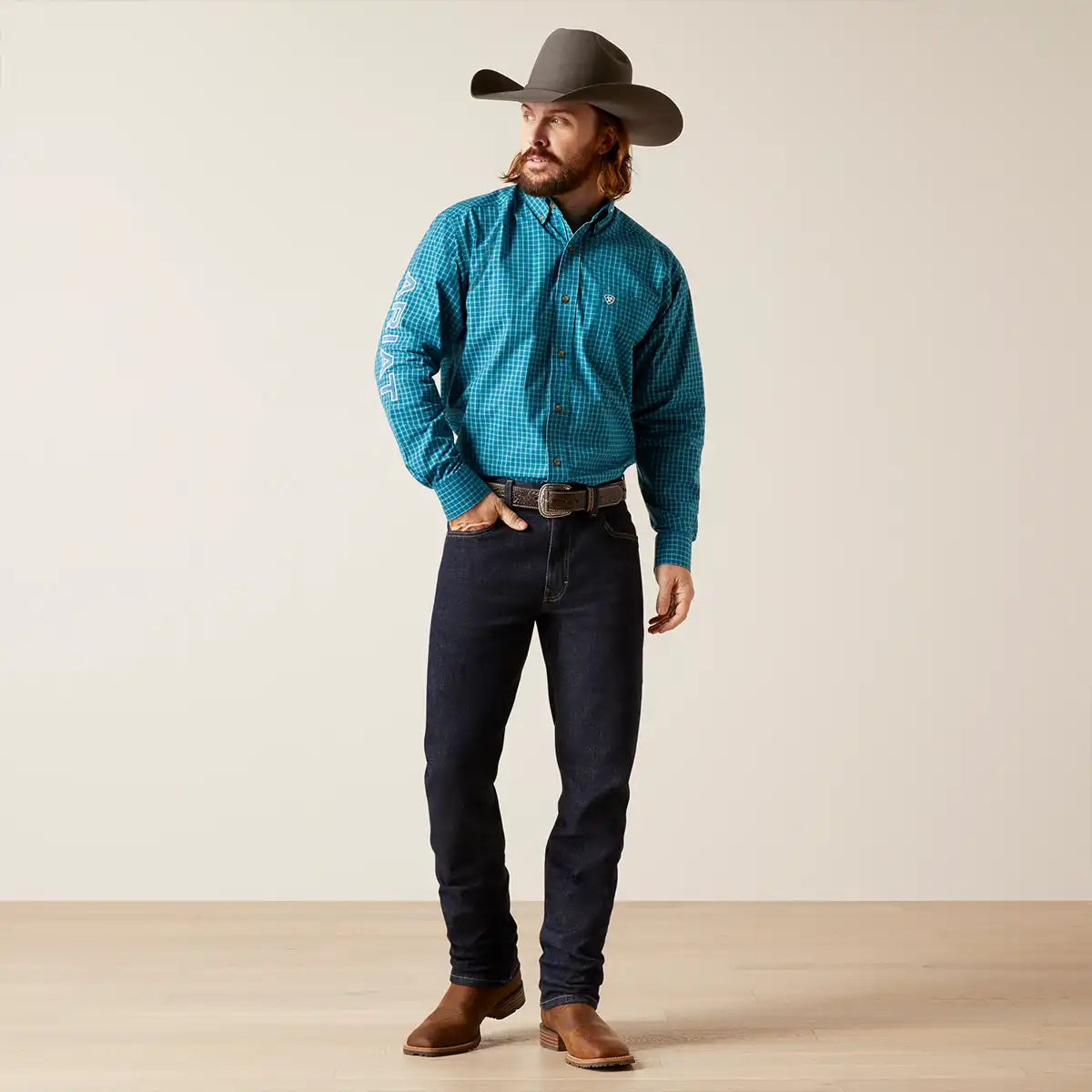 A man wearing a turquoise and black checkered long-sleeve western Ariat shirt, dark blue jeans, brown boots, and a brown cowboy hat, standing against a plain beige studio background.