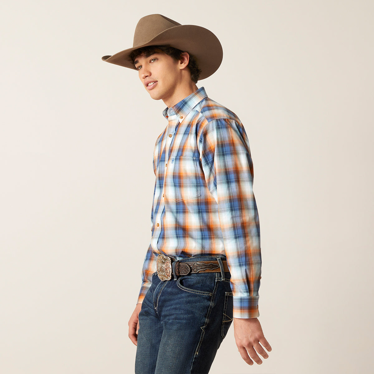 A young man wearing a brown cowboy hat, a blue, orange, and white plaid western long-sleeve button-down Ariat shirt, and dark blue jeans, shown in profile against a light beige studio background.