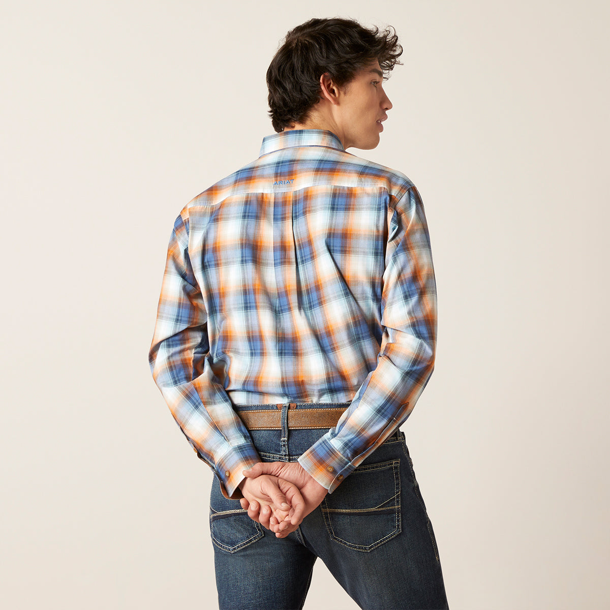 A man wearing a long-sleeve plaid button-down shirt in blue, orange, and white, paired with dark blue jeans, shown from a back-three-quarter view against a plain light beige studio background.