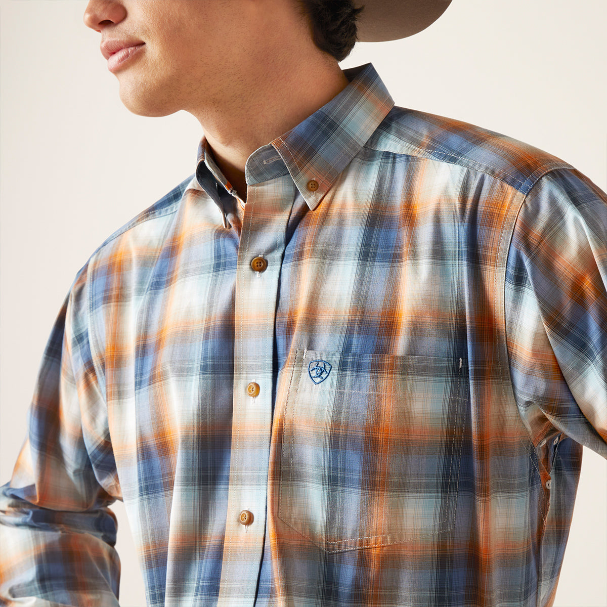A close-up of a man wearing a long-sleeve plaid button-down shirt in blue, orange, and white, with a small embroidered logo on the chest pocket.