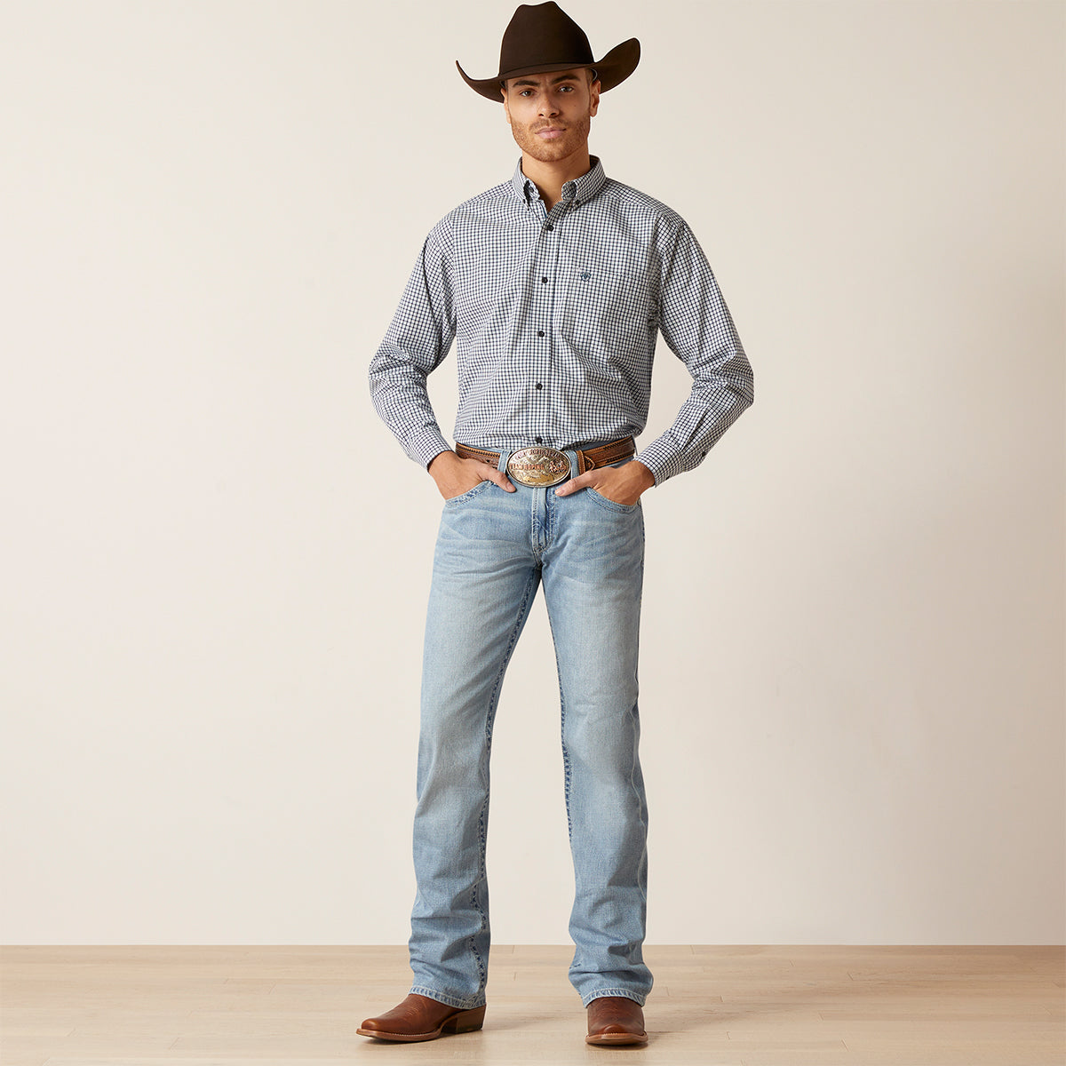 A man modeling a western-style outfit consisting of a light blue and white checkered long-sleeve button-down Ariat shirt, light-wash blue jeans, a brown cowboy hat, and brown leather boots, standing against a plain beige studio background.