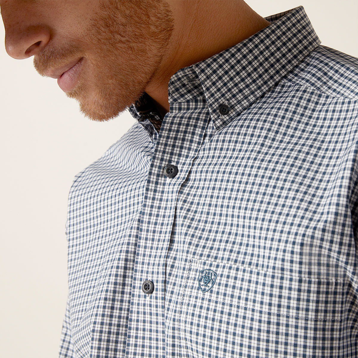 Man wearing a blue and white checkered shirt with a visible Ariat brand logo on a light background.