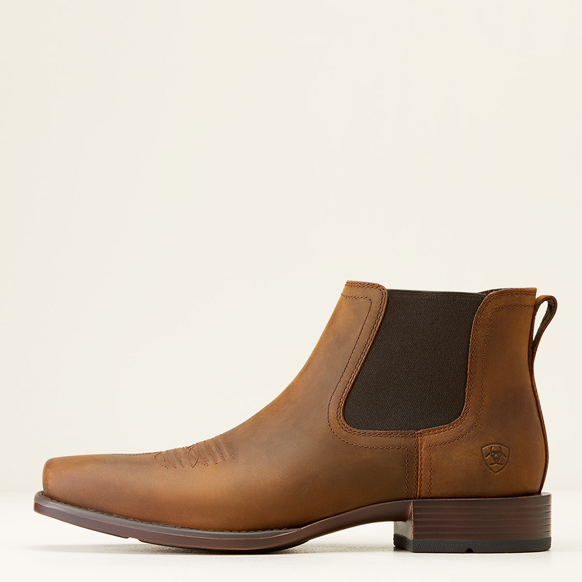 A brown leather ankle Ariat boot with a dark brown elastic side panel, shown in profile against a light cream background. The boot features a low block heel and a small shield logo on the heel.