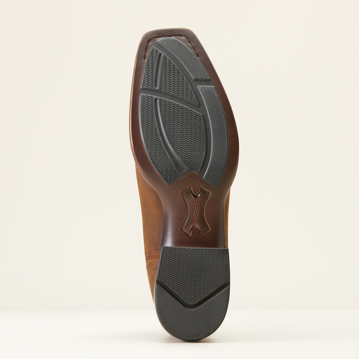 The Ariat outsole of a brown leather boot, showing a textured dark gray tread pattern and a debossed logo on the heel, photographed against a light beige background.