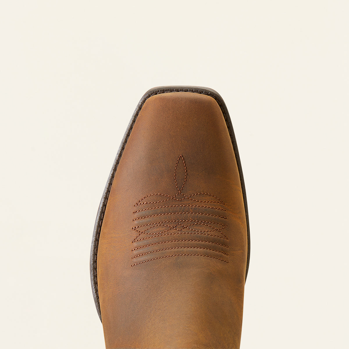 A close-up top-down view of a brown leather Ariat square toe showing decorative stitching and a square edge, set against a solid off-white background.