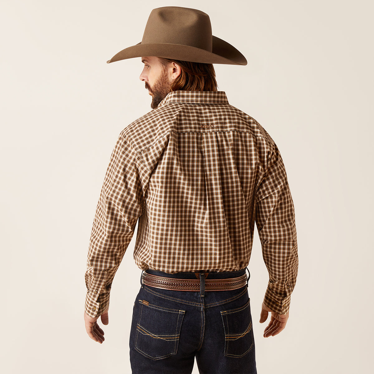A man wearing a brown and tan checkered western Ariat shirt, a brown cowboy hat, and dark blue jeans, shown from the back against a light beige studio background.