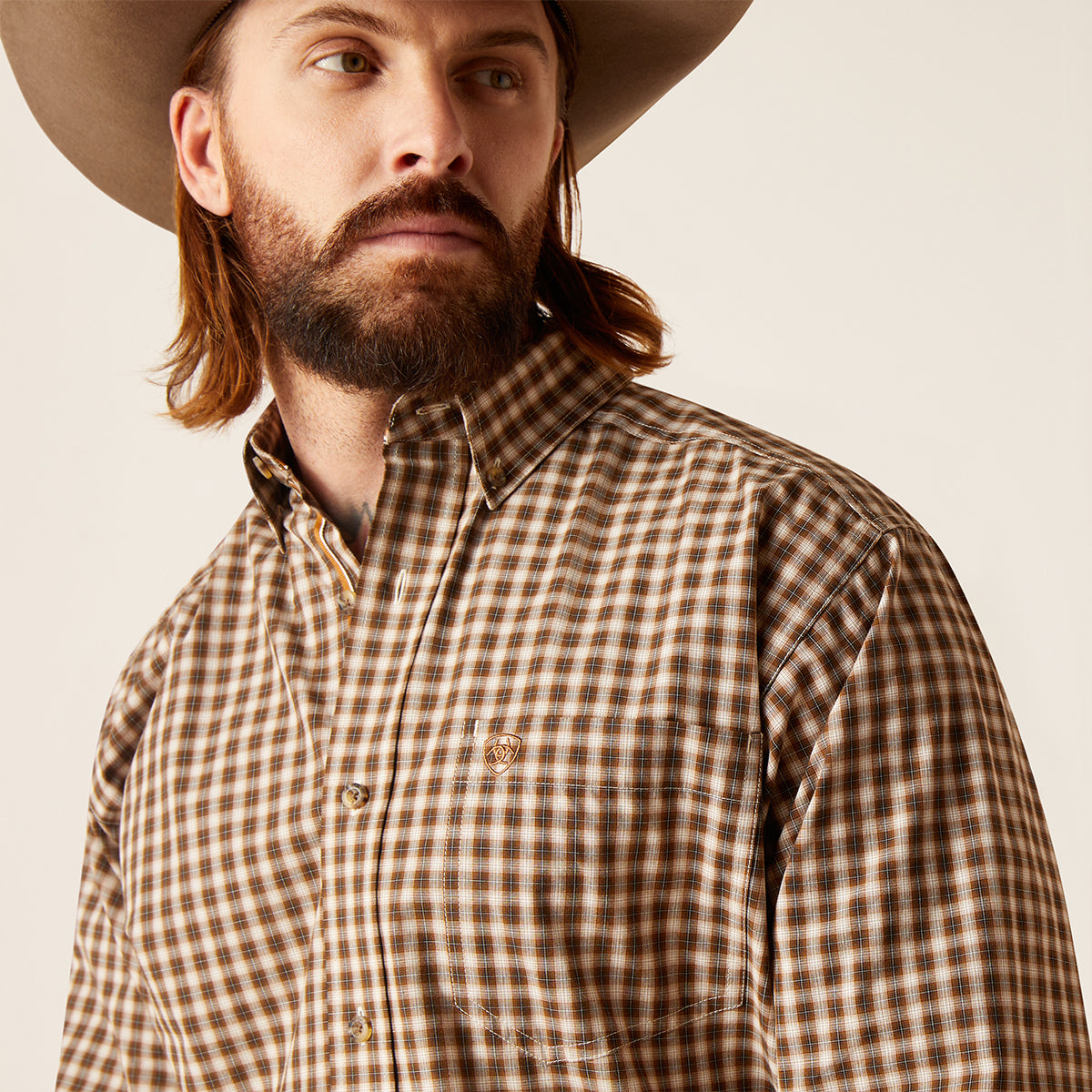 A close-up of a man wearing a brown and tan checkered western-style button-down shirt and a cowboy hat, photographed against a plain light background.