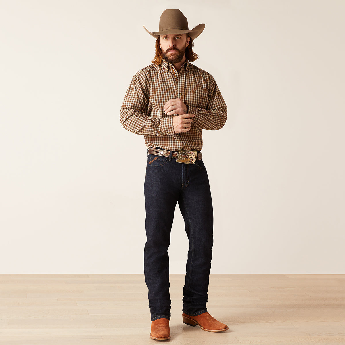 A man wearing a brown and beige checkered long-sleeve button-down Ariat shirt, dark blue jeans, a brown cowboy hat, and brown leather boots, standing against a plain off-white studio background.