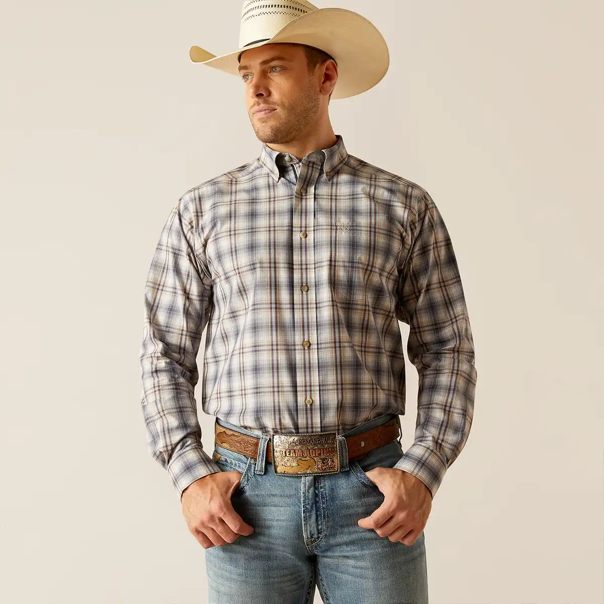 A man wearing a long-sleeve plaid western button-down Ariat shirt in gray, beige, and brown tones, paired with blue jeans, a brown leather belt, and a cream-colored cowboy hat, standing against a solid light beige background.
