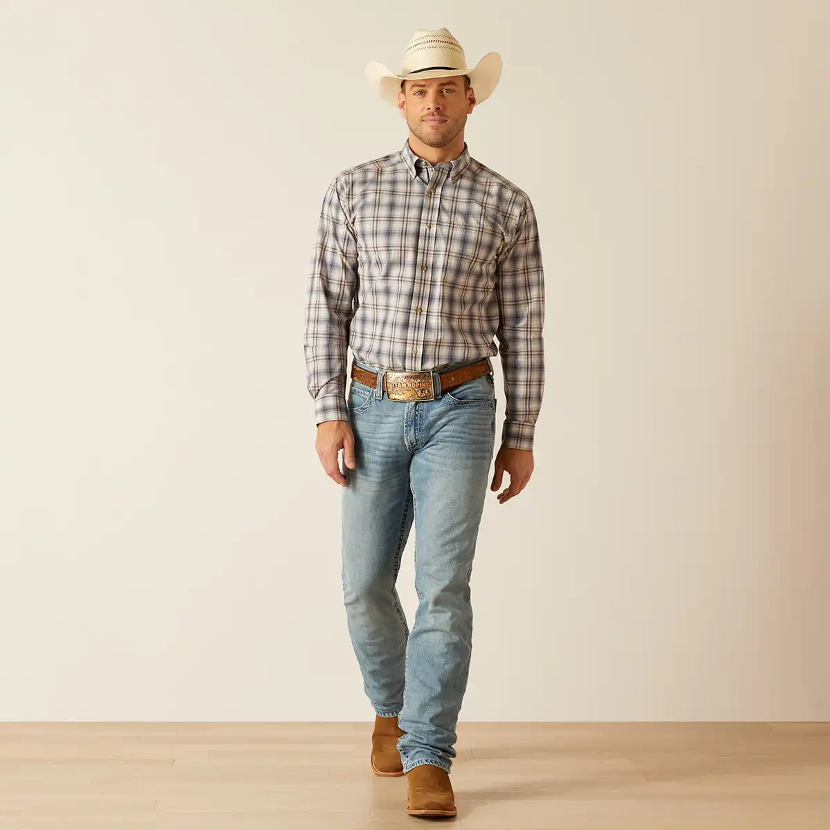 A man wearing a long-sleeve plaid western Ariat shirt, light blue jeans, a brown belt, and a cream cowboy hat, standing against a plain beige studio background.