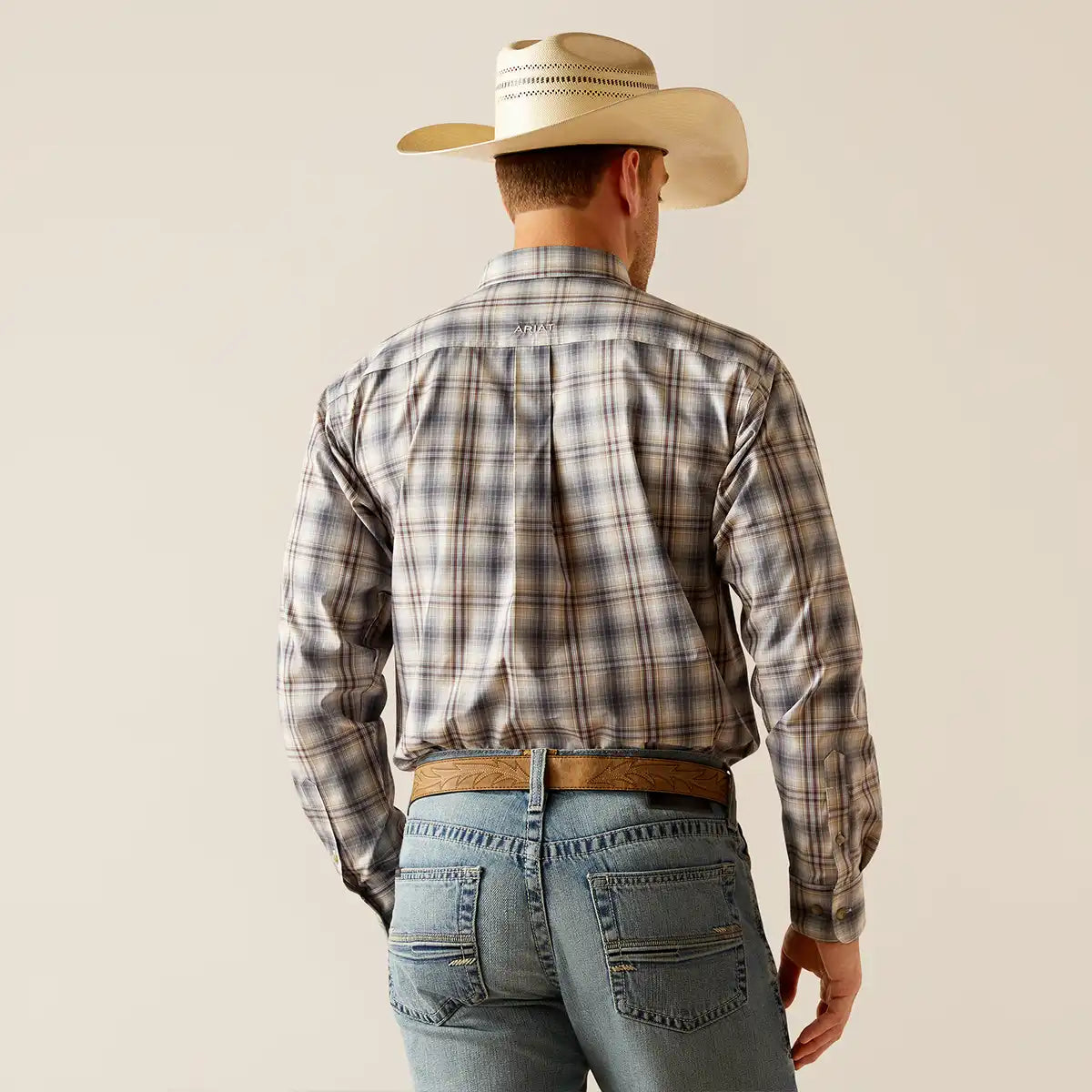 A man viewed from behind wearing a long-sleeve plaid button-up Ariat shirt, a light beige cowboy hat, a brown belt, and blue jeans against a plain light beige studio background.